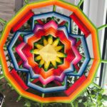 Mandala artesanal multicolor apoyado sobre plantas, con diseño en capas rojas, amarillas y azules.