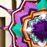 Detalle lateral de un mandala artesanal en tonos morado, verde, blanco y dorado.