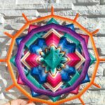 Mandala artesanal multicolor con borde naranja sostenido por una mano frente a una pared de piedra clara.