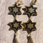 Pendientes artesanales con diseño de estrella en chaquiras Miyuki negro y dorado, con borla decorativa.